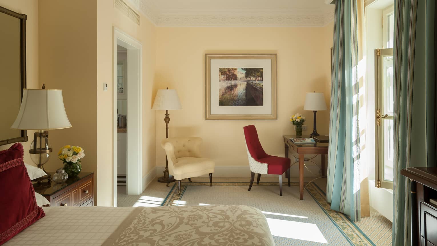 Deluxe Room bed, white armchair, red chair at desk by sunny window with curtains