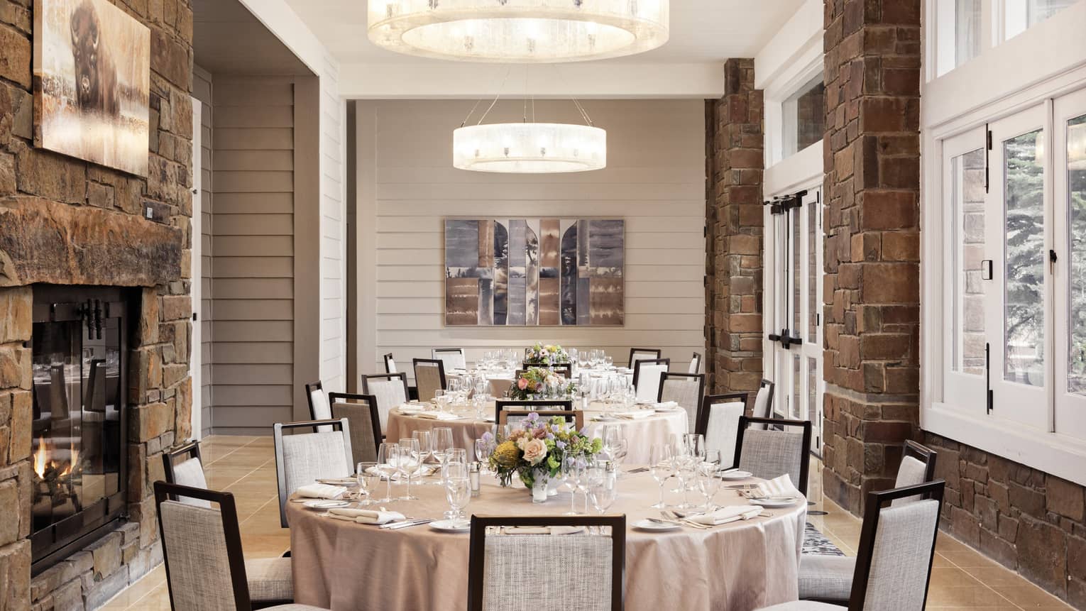 A row of round tables set for a wedding, featuring floral centrepieces, cushioned chairs, a stone fireplace and large windows 