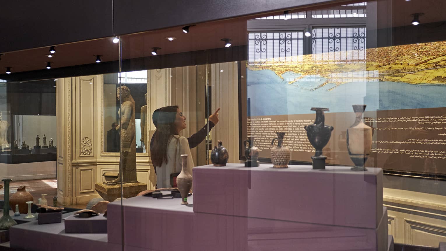 A visitor in a museum points up at a historical map lit up on a wall, behind them a display case with various vases and jugs.