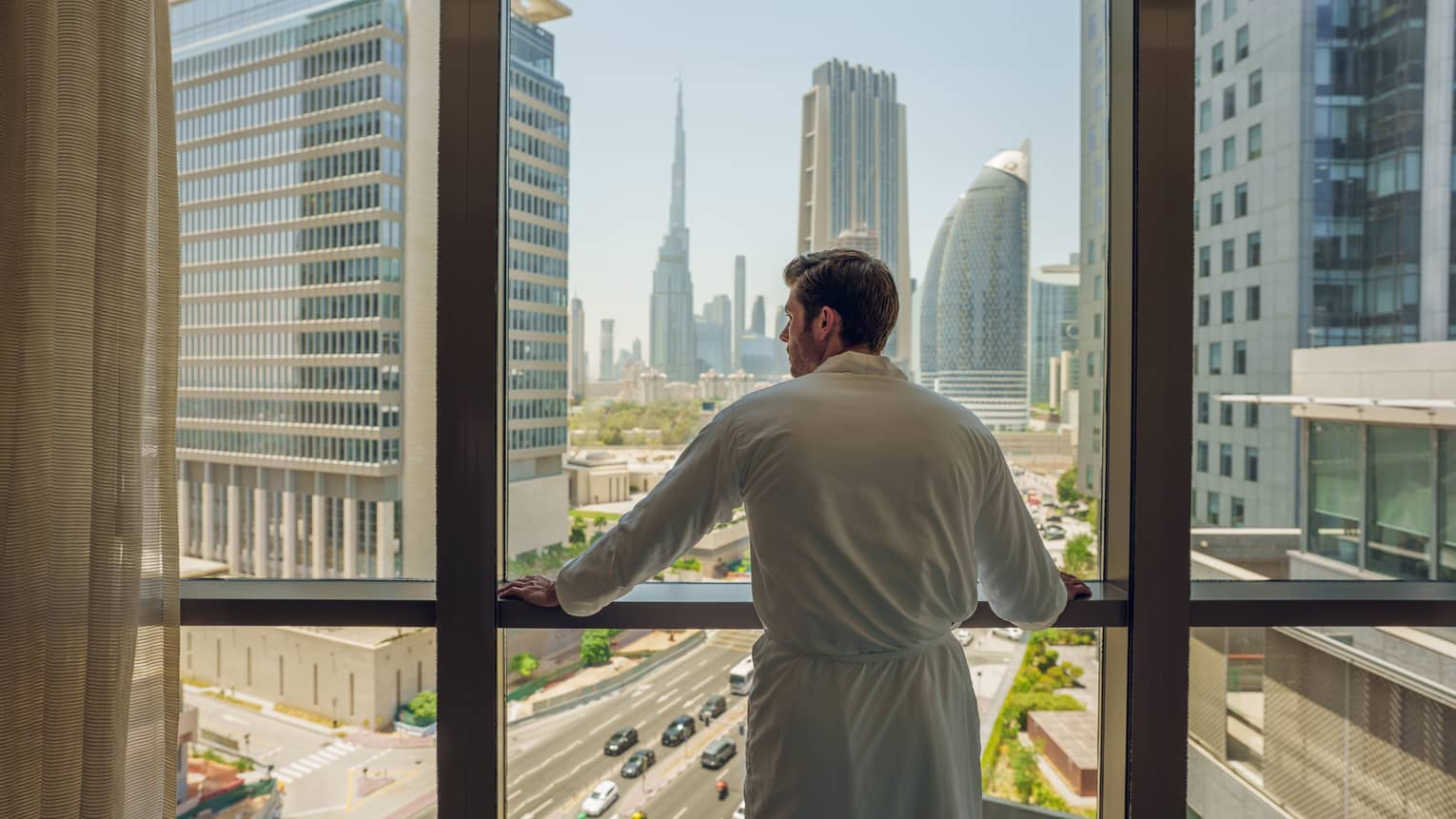 Man wearing a white spa robe stands at a window overlooking a city skyline