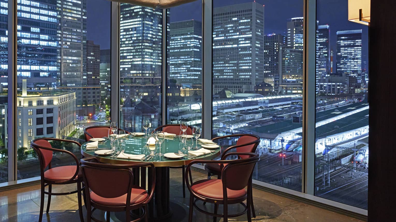 Luxury boutique hotel dining setup with a round table elegantly set for guests, offering a panoramic city view at night through tall glass windows
