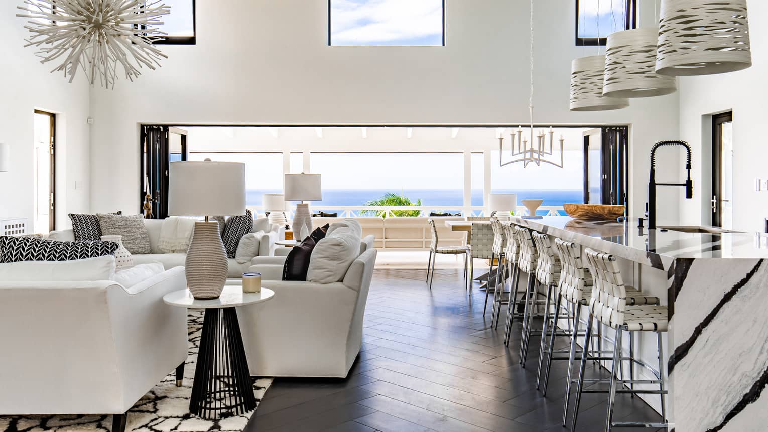 Large common area with long marble kitchen island and bar chairs, white living room furniture, sea view