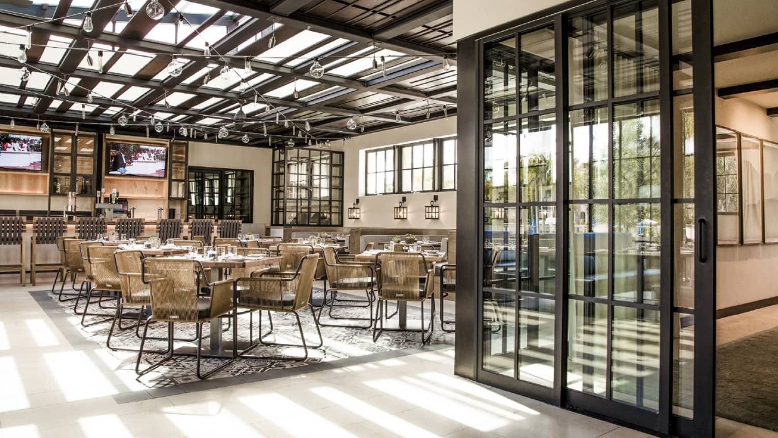 Dining room and bar with glass roof, modern wicker chairs and tables, glass sliding doors with black frames, glass bulb light strings