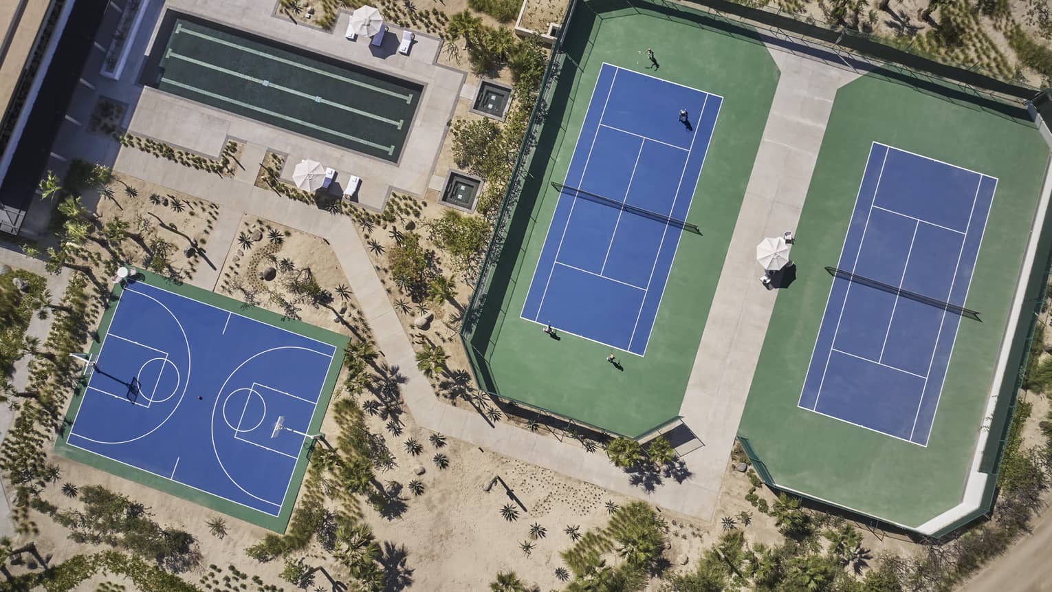 Aerial view of two green and blue tennis courts, one long rectangular pool and one blue basketball court surrounded by sand and greenery