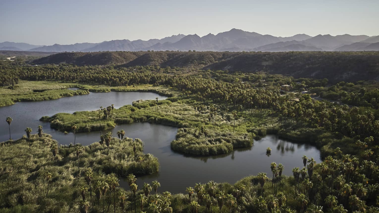 Aerial view of water surrounded by lush vegetation bordered by hills, with distant mountains visible under a calm ombre sky.