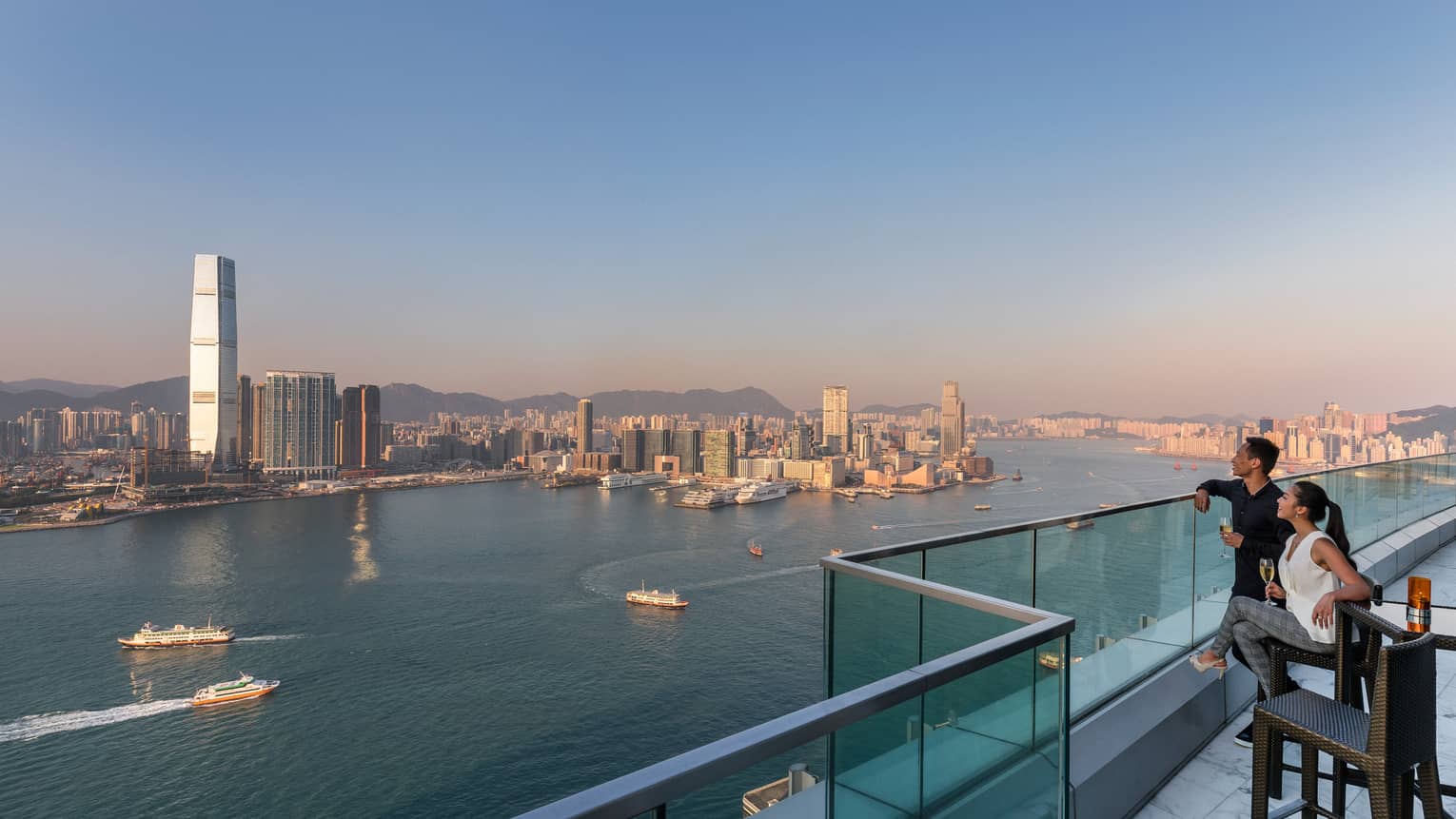 Two people standing on a rooftop terrace, enjoying a panoramic view of a harbor with boats and a city skyline in the distance during the day.