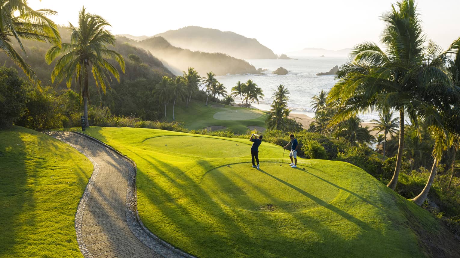 Rear view of two golfers on an elevated tee, one swinging toward the green down by the beach, misty hills and ocean beyond.