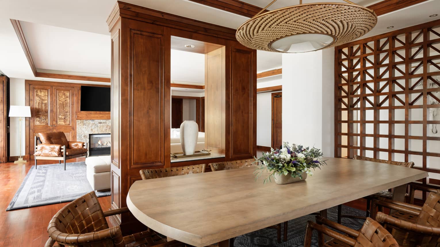 Luxury suite dining area with a large wooden table, woven chairs and a floral centrepiece, with an open view into a cozy lounge with a fireplace