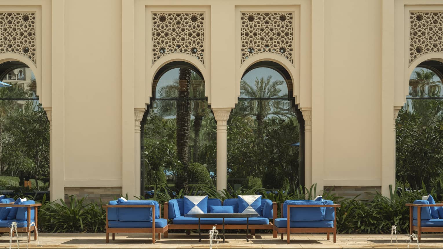 A sunny courtyard with upholstered seating and greenery, with an exterior hotel wall with tall, elegant arched windows in the background