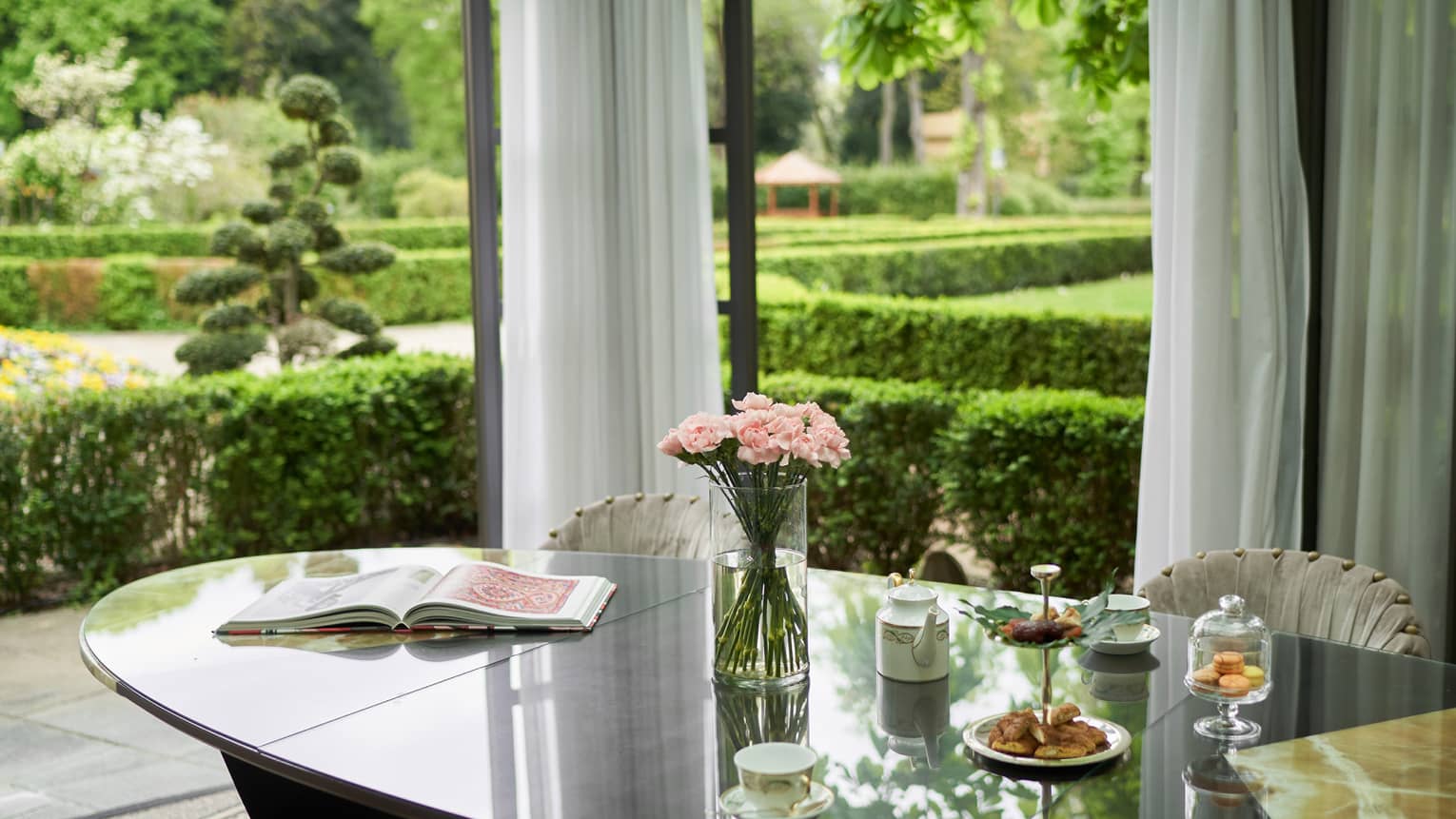 Oval dining table set with afternoon tea and flowers, garden in the background