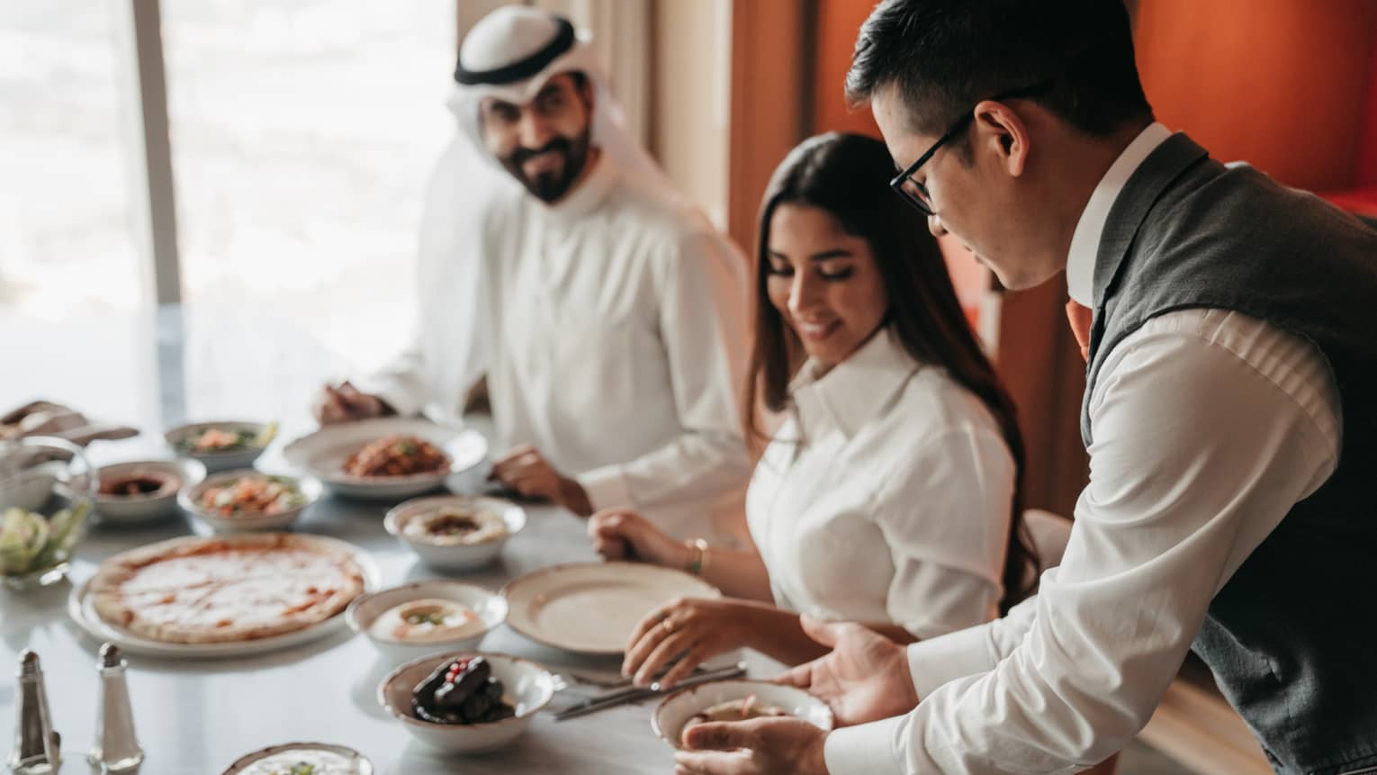 A couple dressed in white, being served a full meal by a staff member.