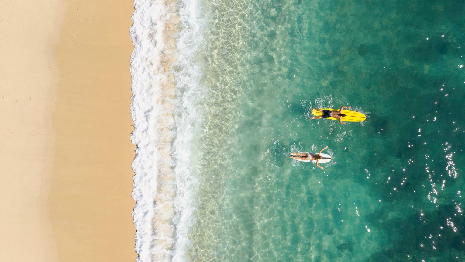 Bird’s-eye view of two people paddling their boards on their stomachs as frothy waves break on the beach behind them.
