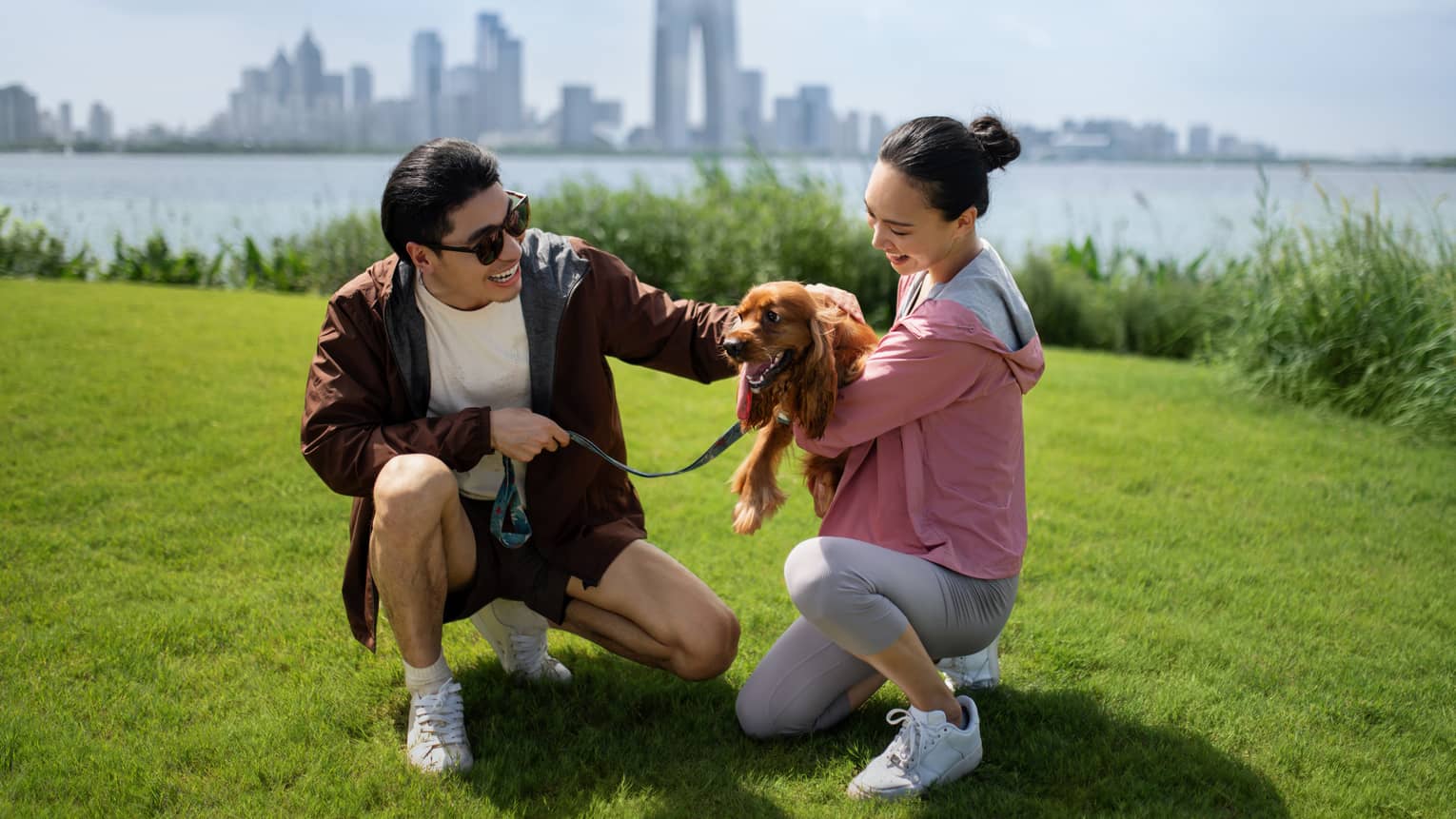 A laughing couple crouch to cuddle a long-eared hound on a grassy shoreline, calm water and distant skyscrapers behind them.