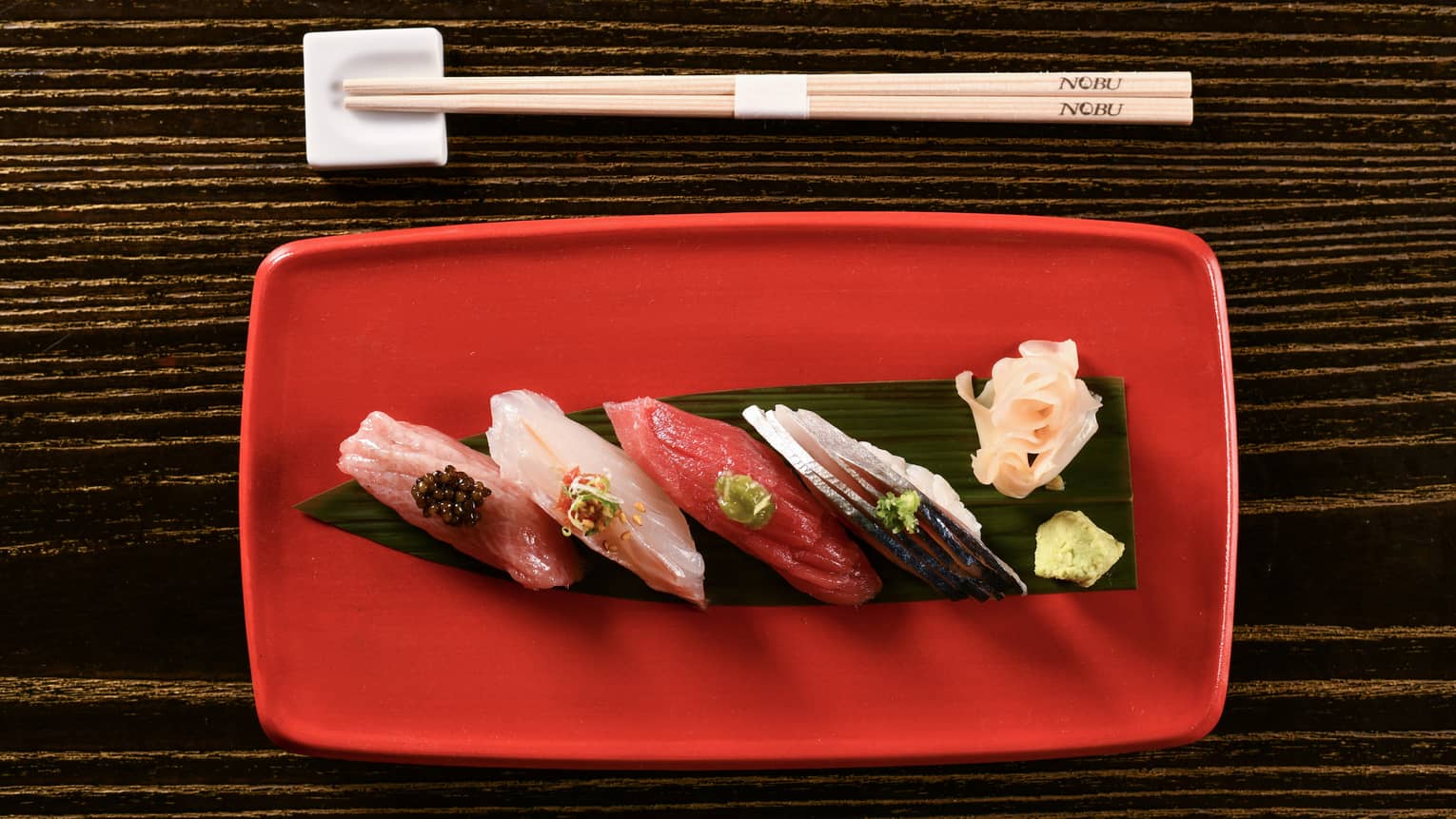 Four different pieces of sashimi, each with a small elegant garnish, rest on a red plate below Nobu-branded chopsticks.