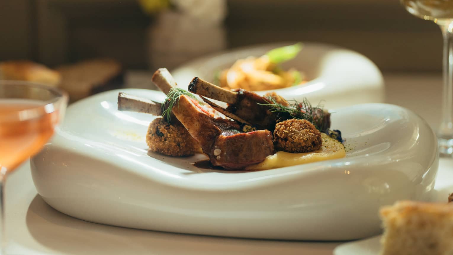 Lamb chops plated on a thick, white organic-edge dish and garnished with sprigs of greenery