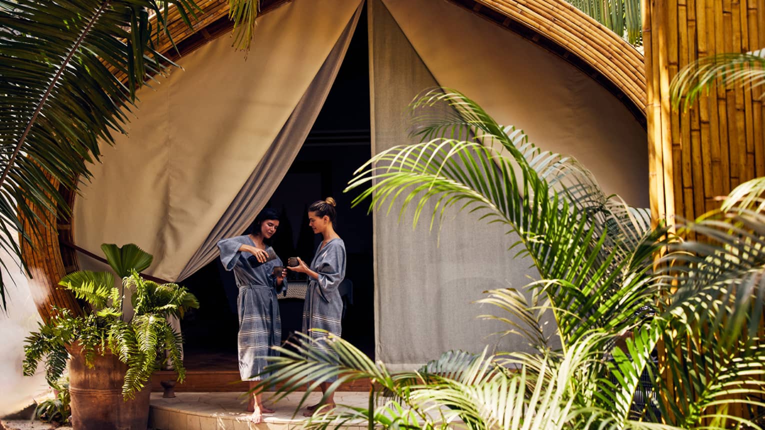 Two women in spa robes stand outside a luxury spa tent, one pouring tea for the other, surrounded by lush greenery in a peaceful setting.
