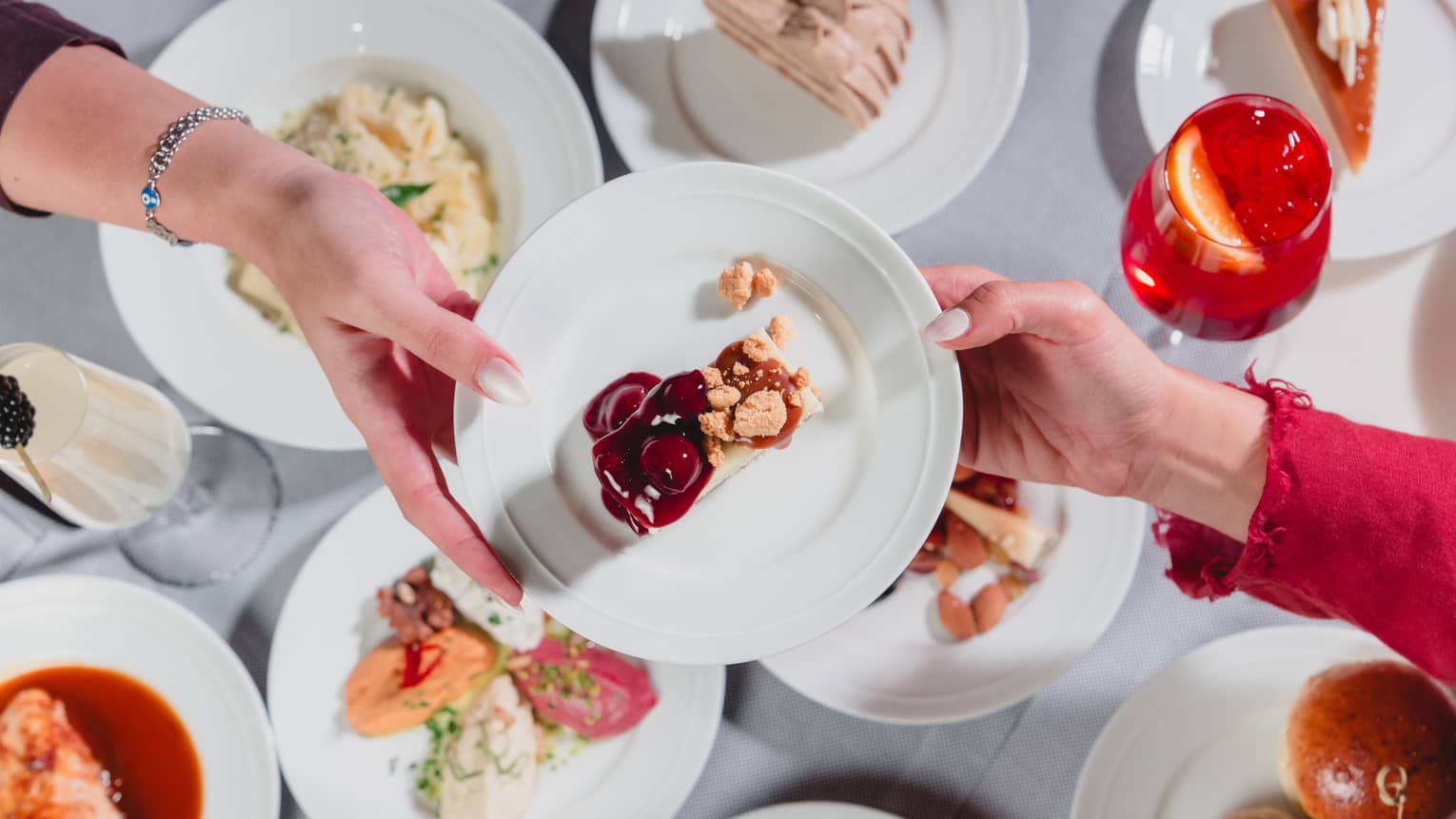 One hand passes along a white plate to another hand with a variety of dishes of food seen below on a table