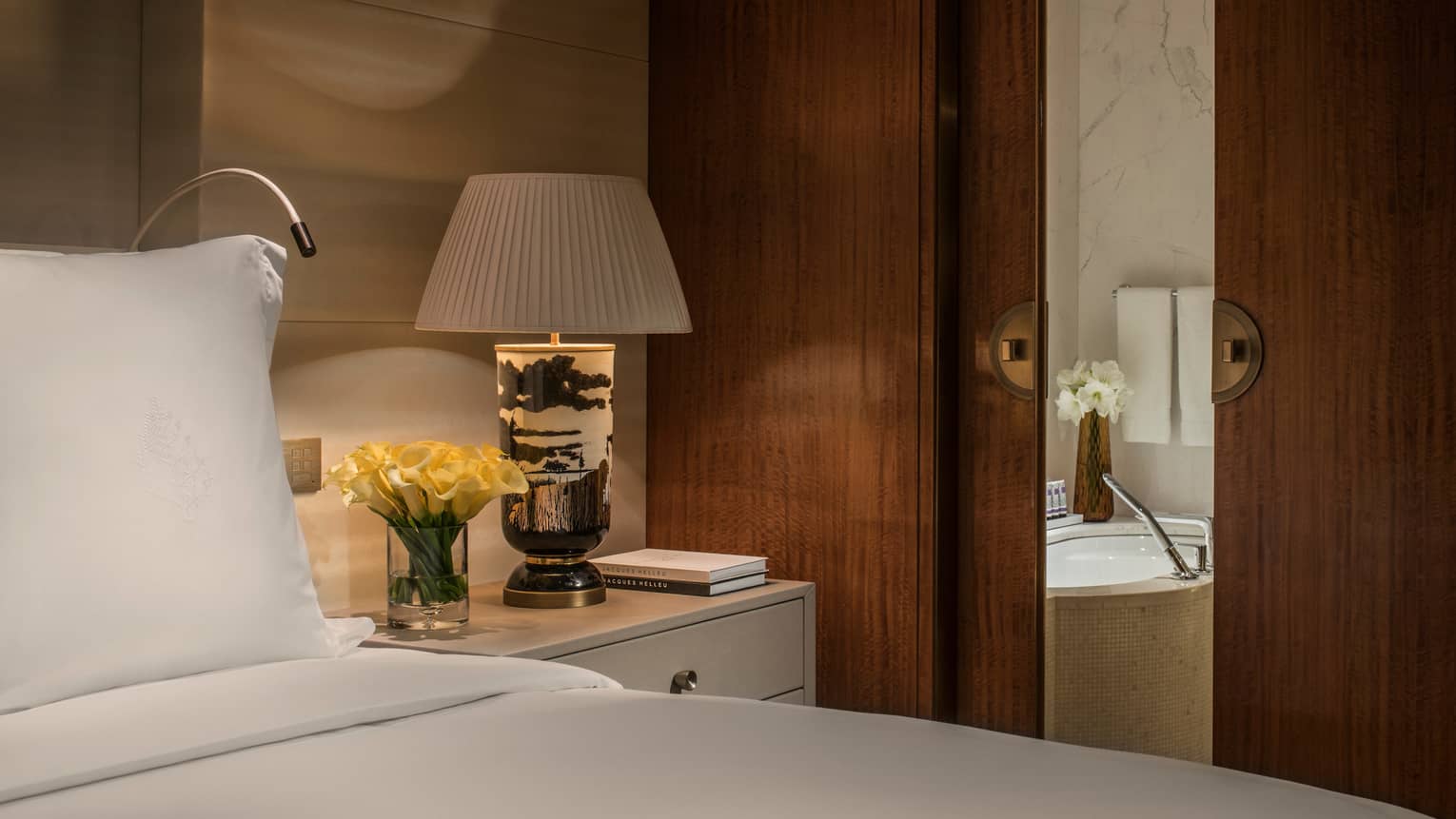 Club Four Seasons Room close-up of bed pillow, nightstand with lamp, flowers, wood bathroom door