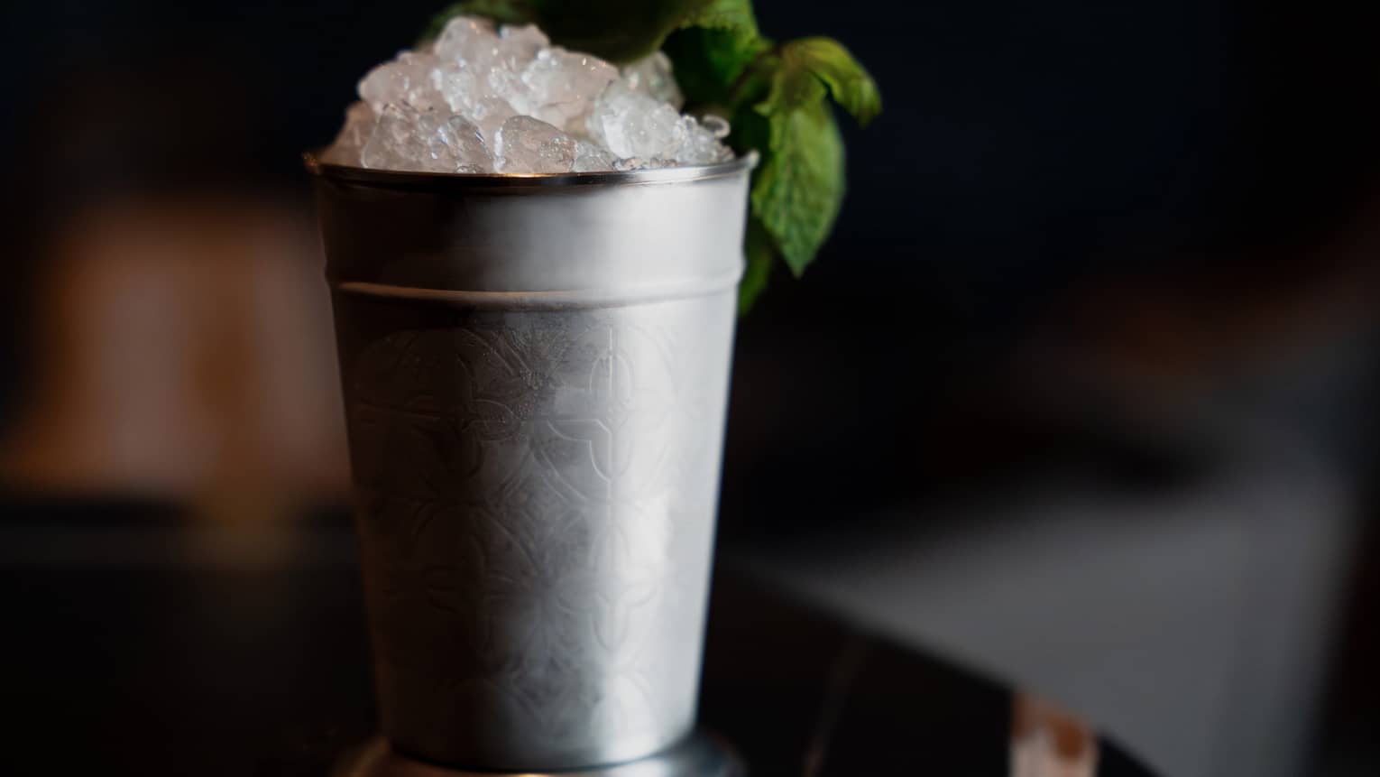 A tall metal tumbler holds a drink topped with ice and garnished with a sprig of green leaves.