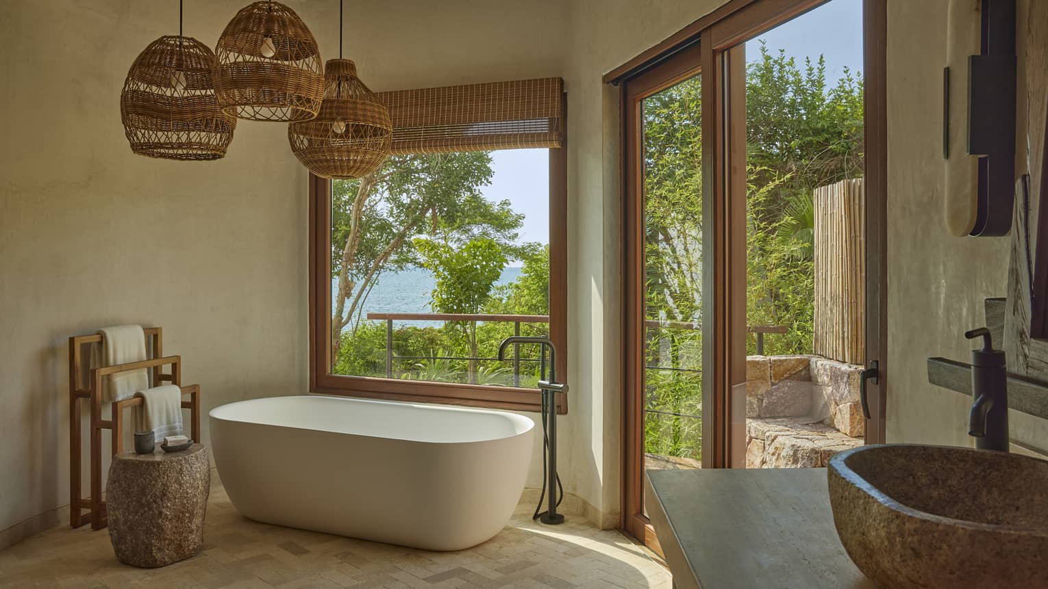 Bathroom with freestanding tub, three hanging whicker lights and window to tropical forest