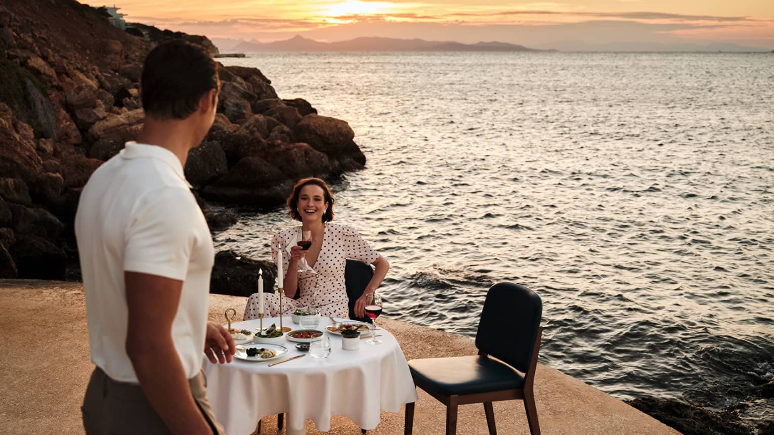 A person sits at an elegant dining table in an outdoor setting, while another person walks towards the table. A rocky shoreline, the setting sun and the ocean are in the background.