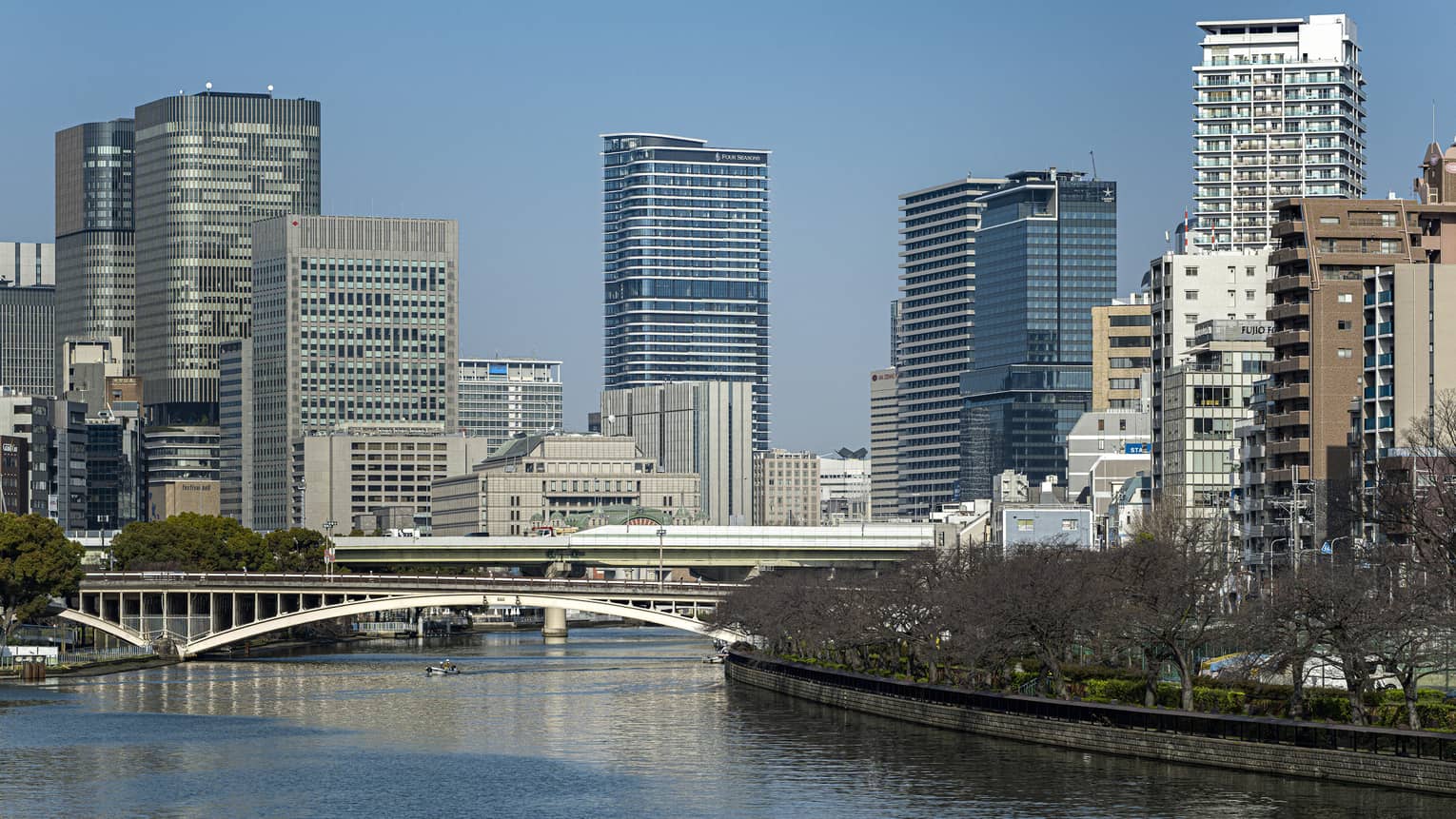 Under a clear blue sky, Osaka's modern high-rises loom above two bridges spanning a river lined with dormant cherry blossoms.