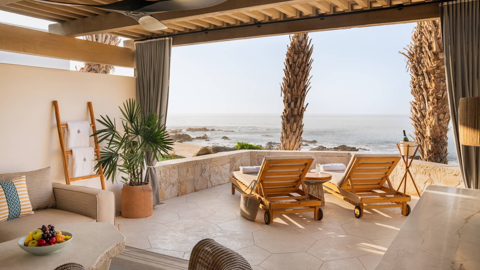 View from inside a neutral cabana with lounge chairs by a low stone wall looking out towards a rocky ocean shoreline.