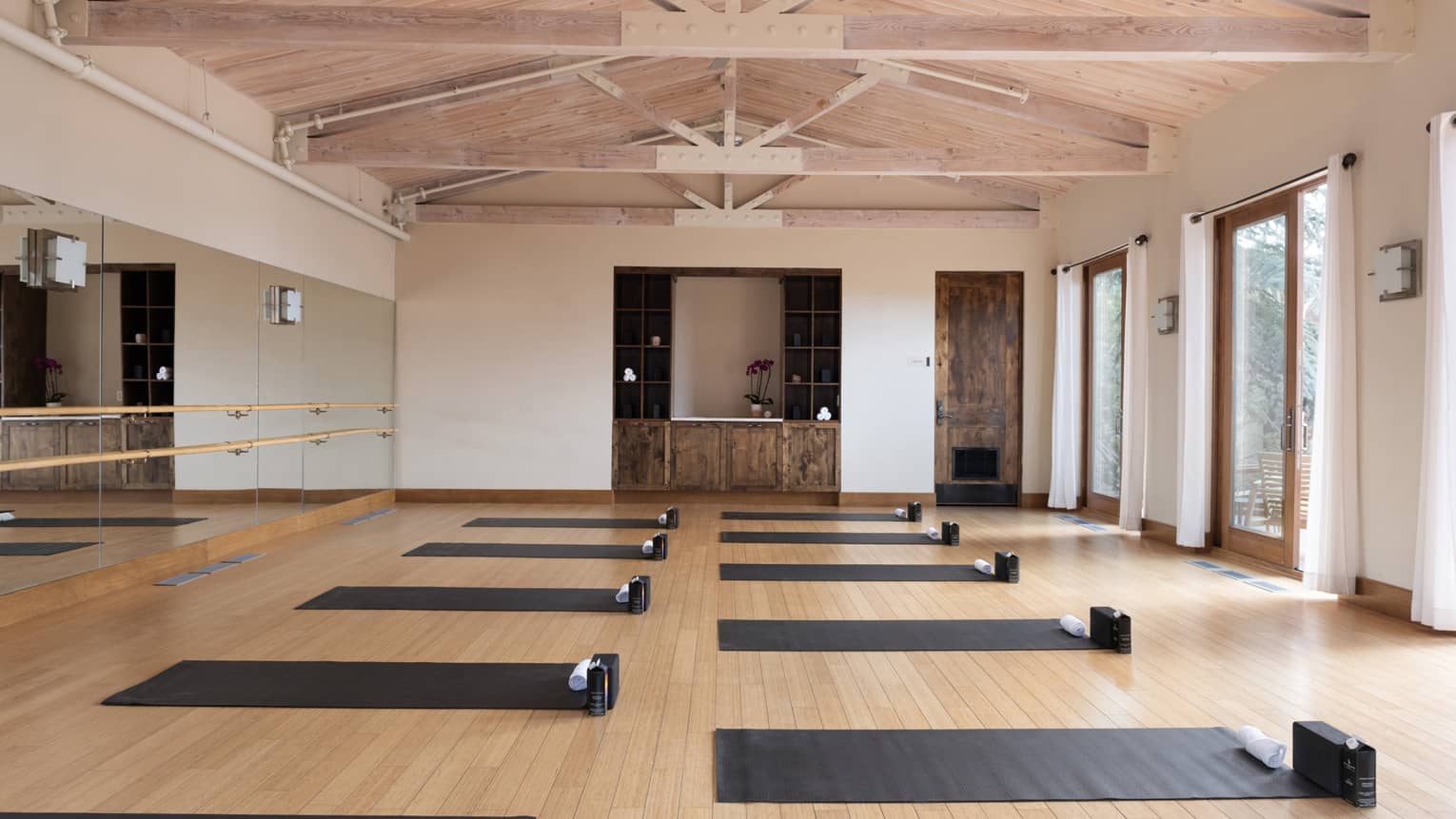 Interior of yoga studio with open rafter ceilings, one mirrored wall and wood floors with two rows of black yoga mats set out on the floor