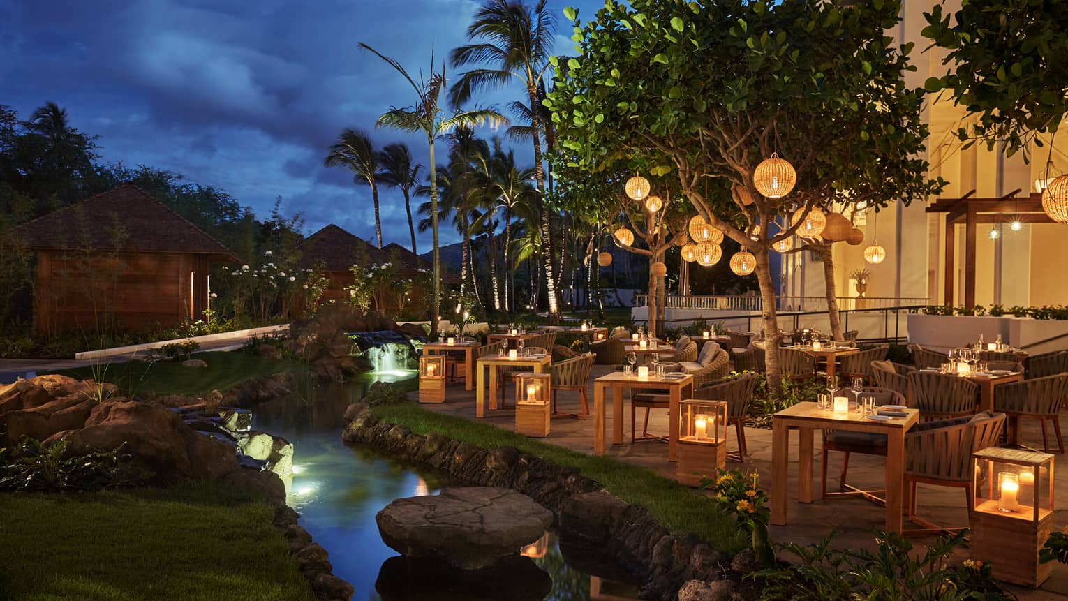 Noe patio dining tables, chairs, lanterns by stream at night
