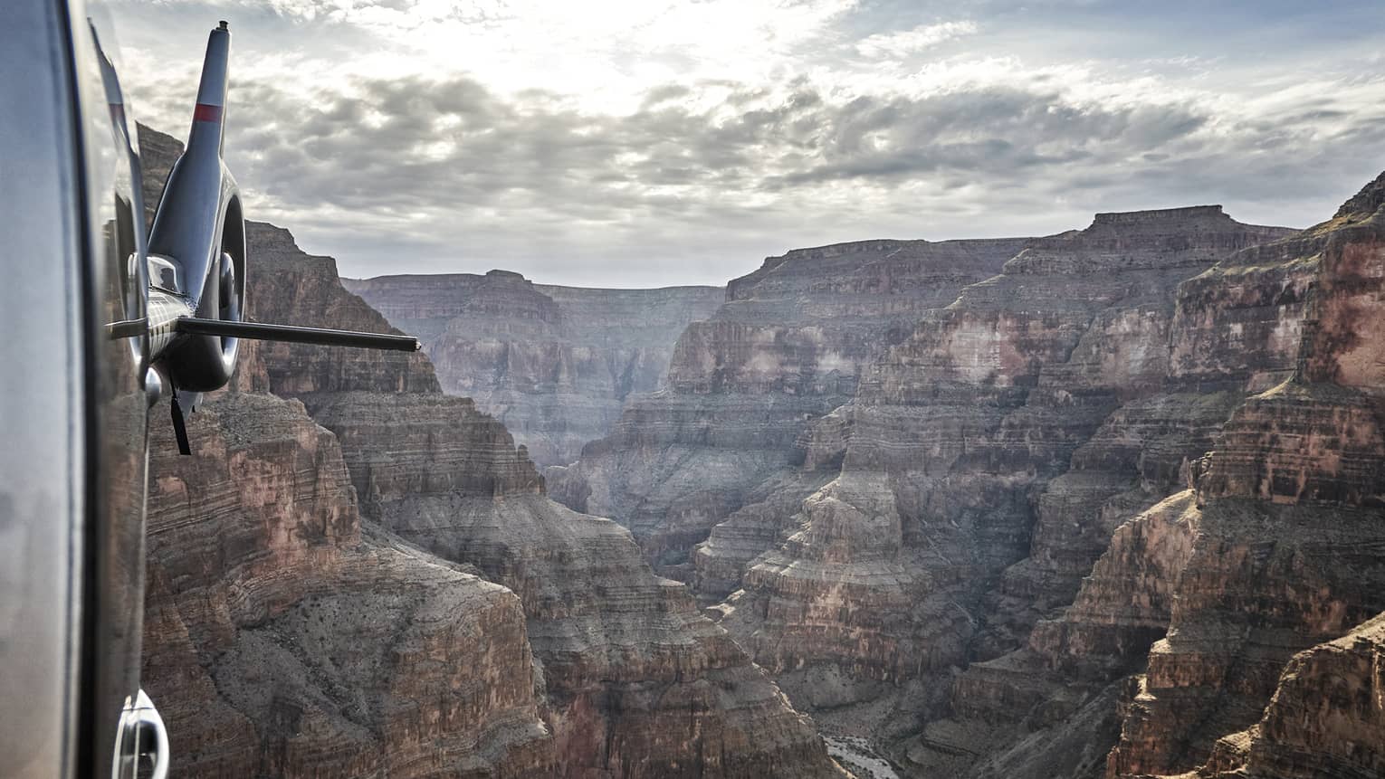 Viewed from a helicopter in flight, an awe-inspiring canyon landscape stretches into the distance beneath a cloudy sky.