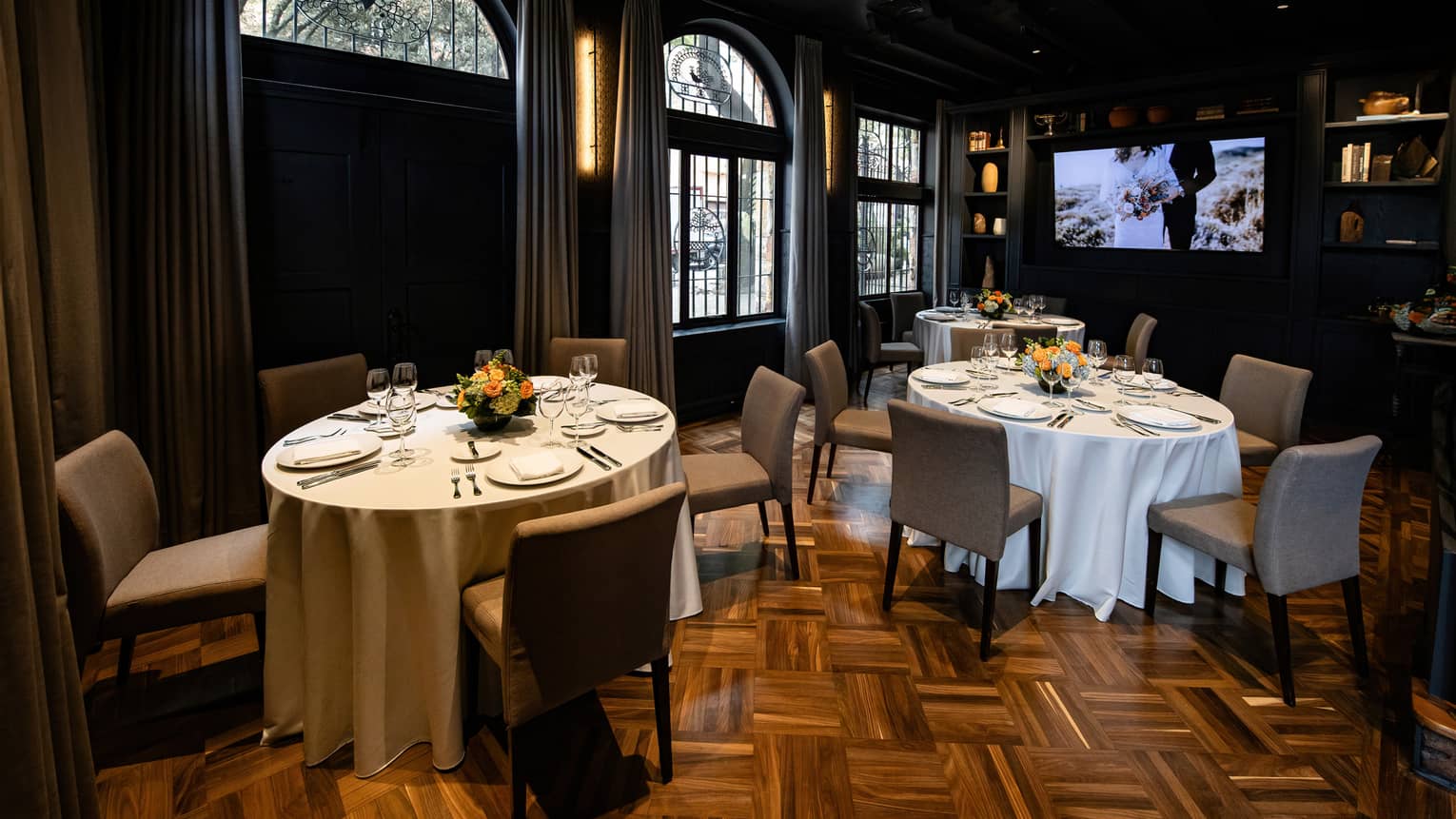 A gently lit, spacious event room with round tables, upholstered chairs, large arched windows, a decorative wood floor and a flat-screen TV. The tables are set for a meal, and wall sconces provide soft light.