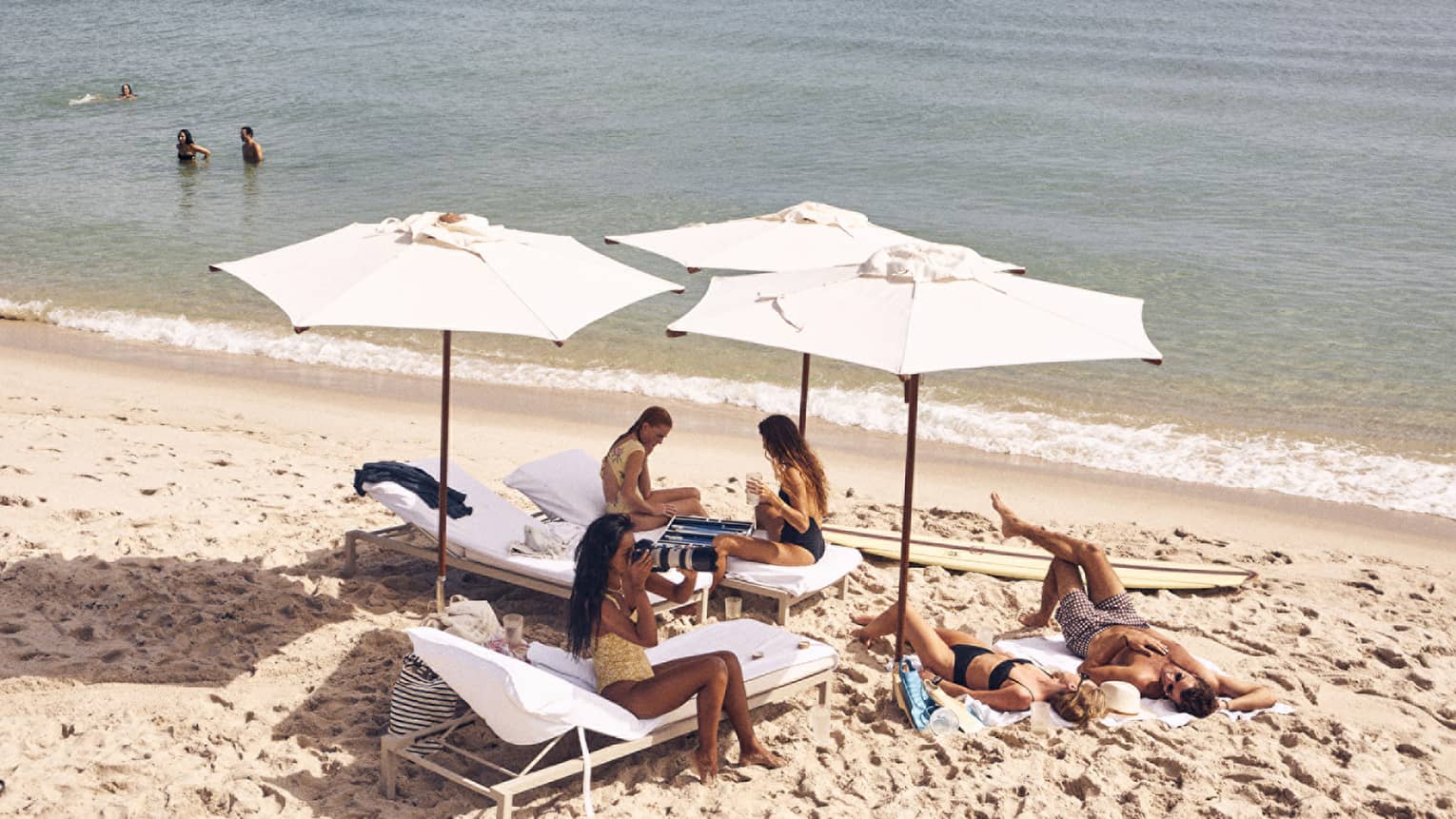 Tourists enjoying a sunny day at the beach by laying on the sand, playing in the water and relaxing under beach umbrellas.