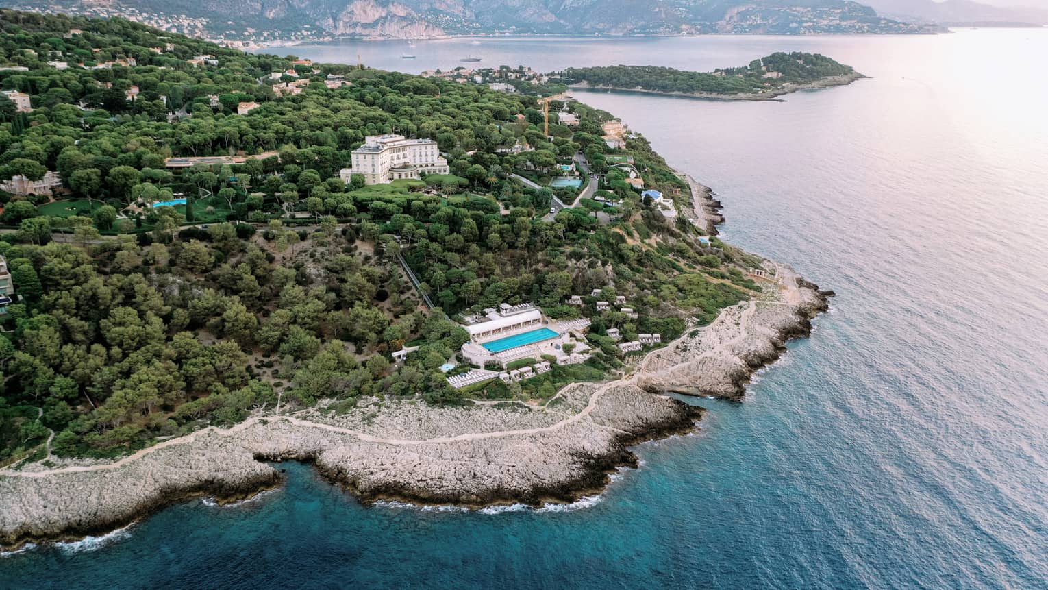 Aerial view of buildings and pools nestled among verdant trees on a sloping hillside along an extensive, rugged coastline.