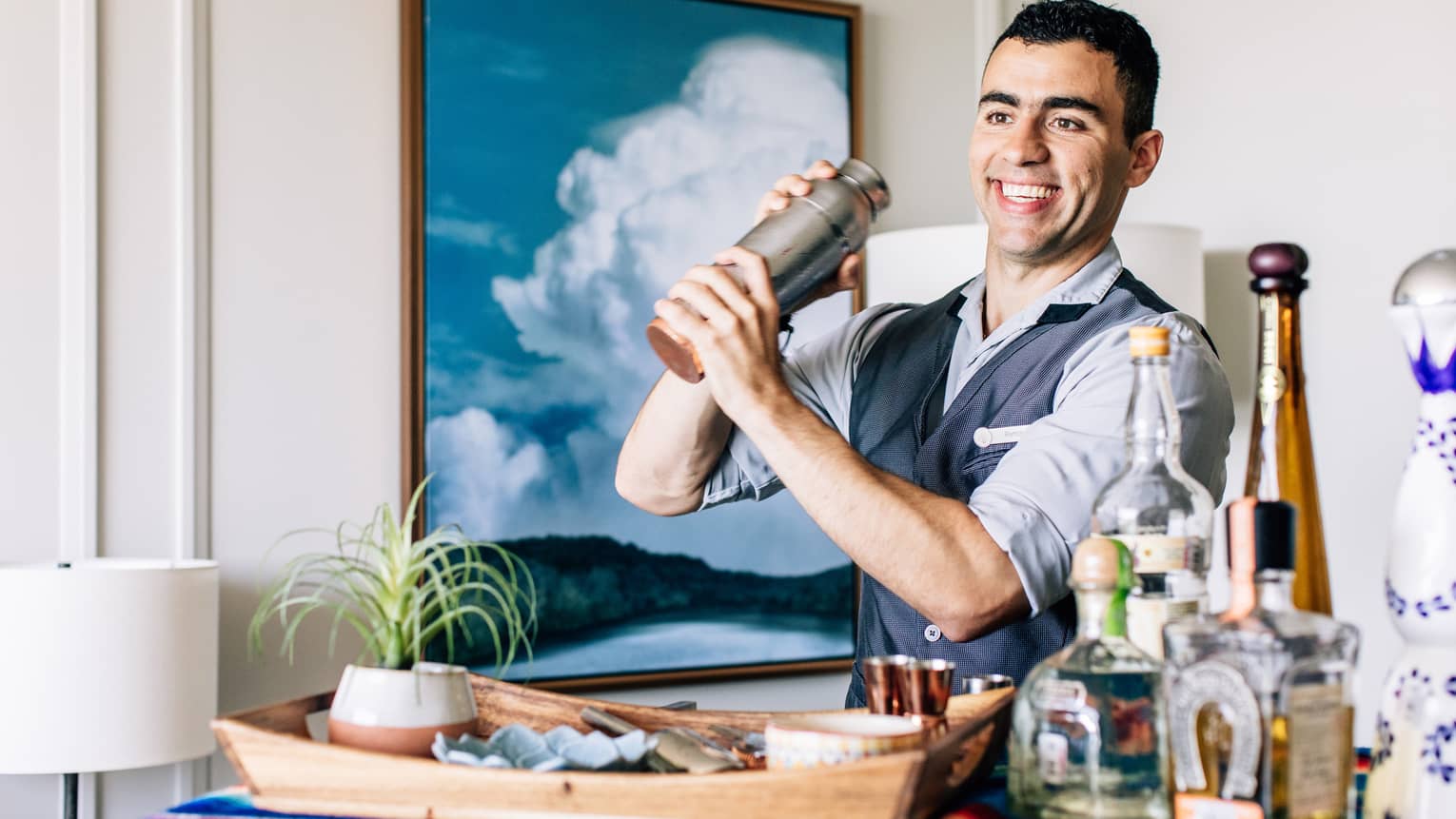 A bartender shaking a cocktail with liquor bottles on a cart in front of him.