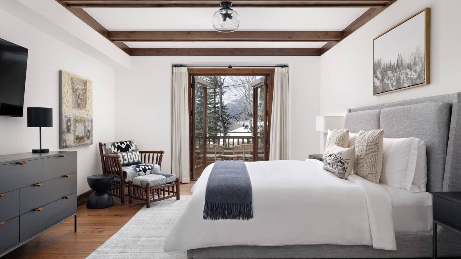 Luxury resort bedroom with a grey upholstered king bed, wall artwork, wooden armchair, dresser with black lamp, and open doors to a snowy forest-view balcony