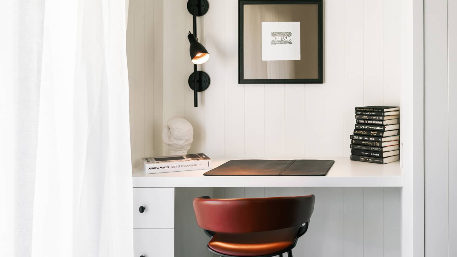 Close-up of a luxury resort room desk with black wall lamp, stacked books, art print, red leather chair and decorative bust on a white workspace