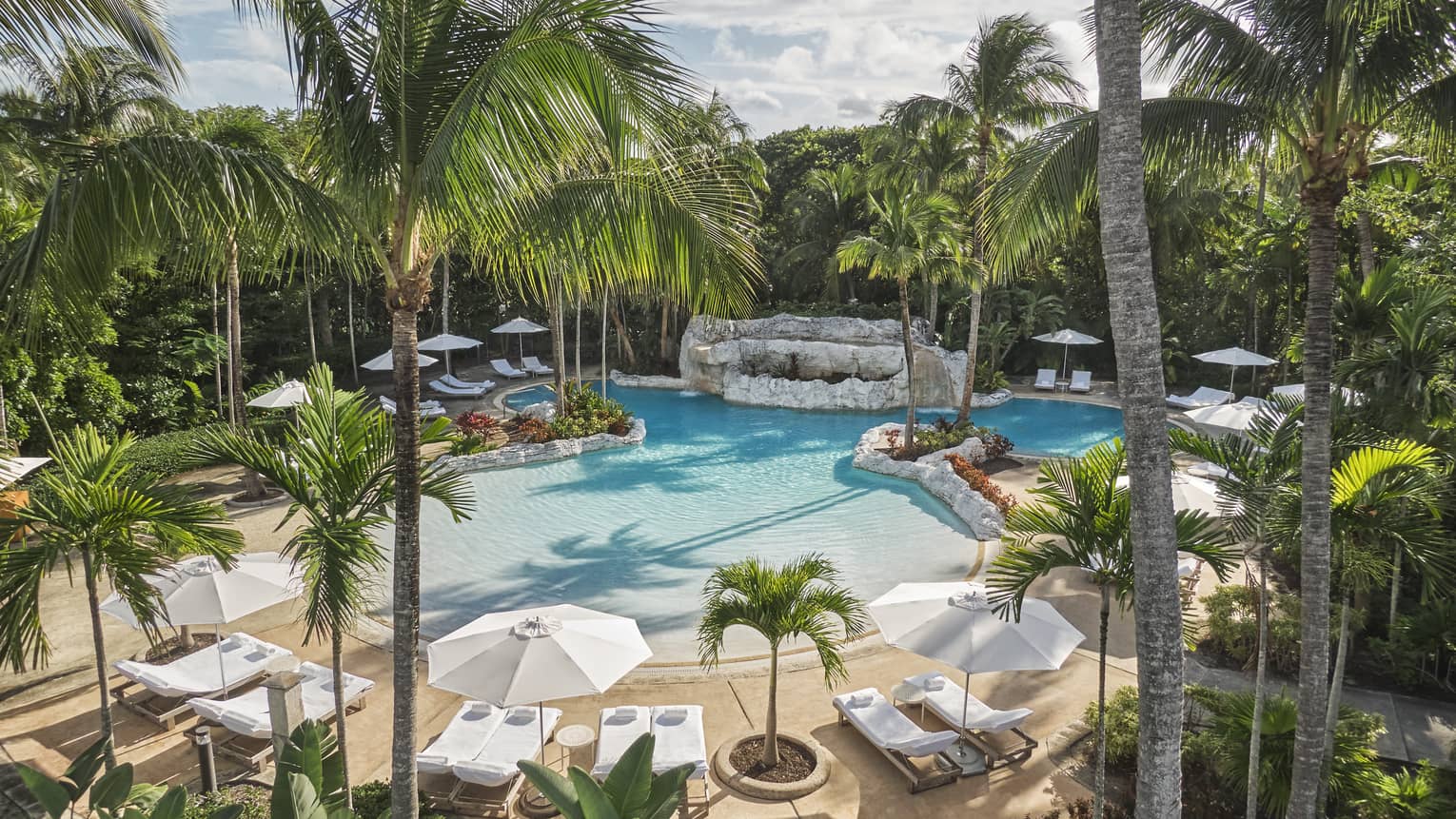 Luxury resort pool surrounded by palm trees, white lounge chairs and large umbrellas