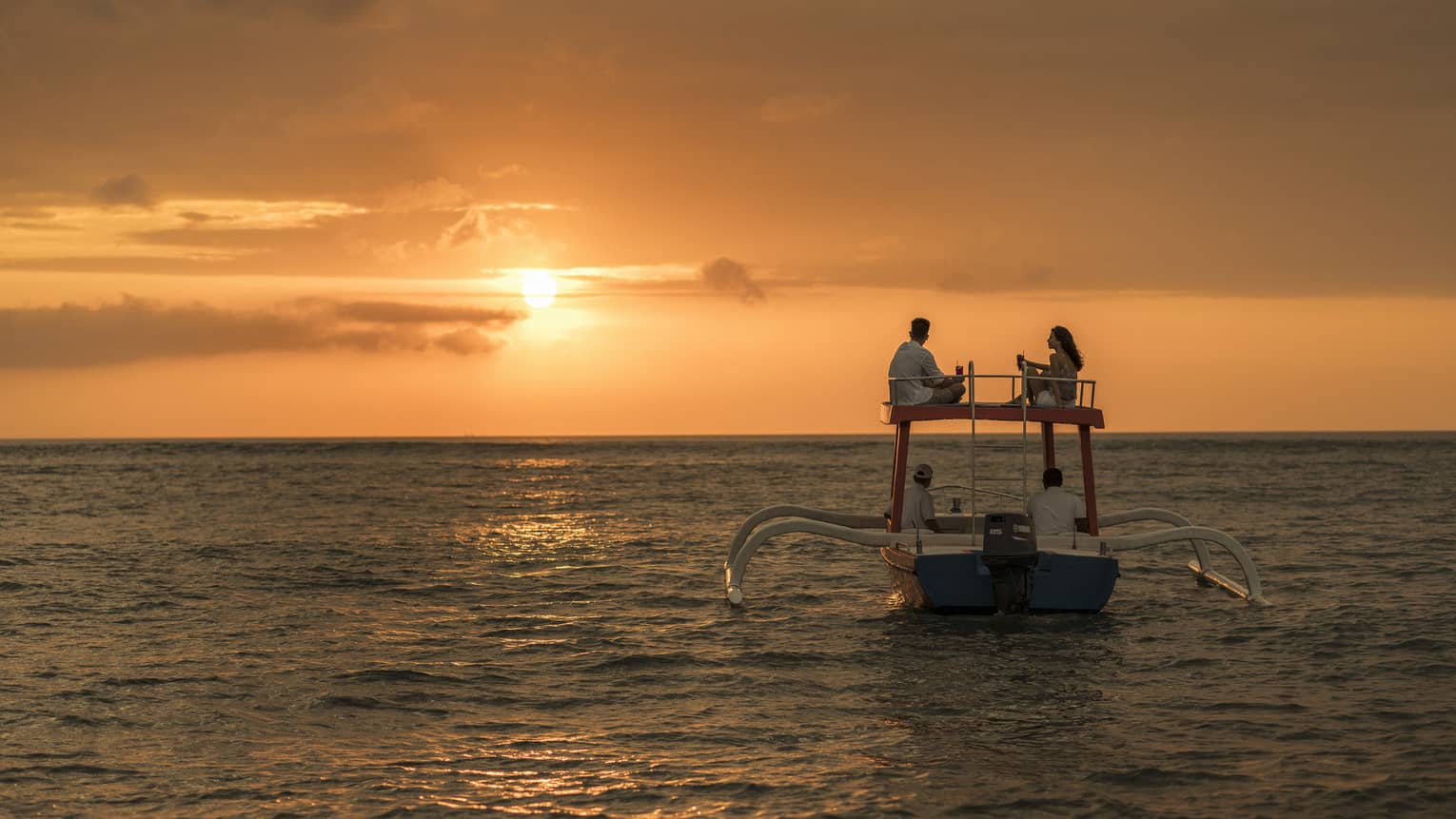 Couple sits on roof of boat sailing on ocean at sunset