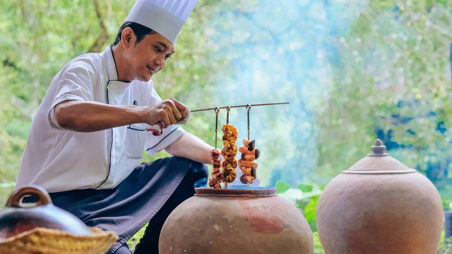 Chef holds prawn skewers over traditional clay cooking pot