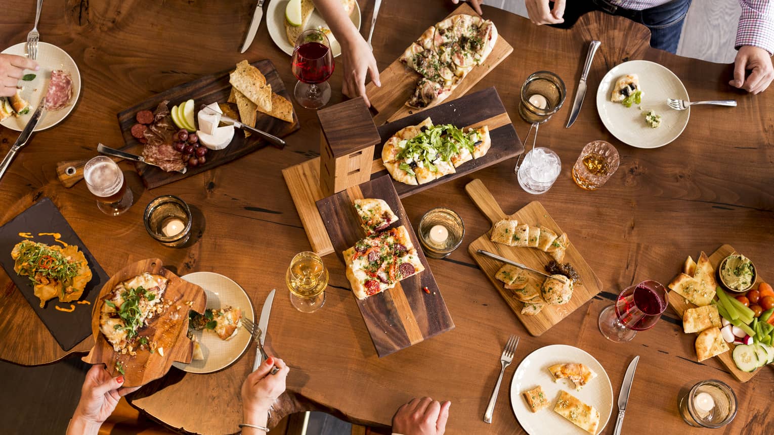 Aerial view of people dining around rustic wood table with platters of flatbreads, appetizers