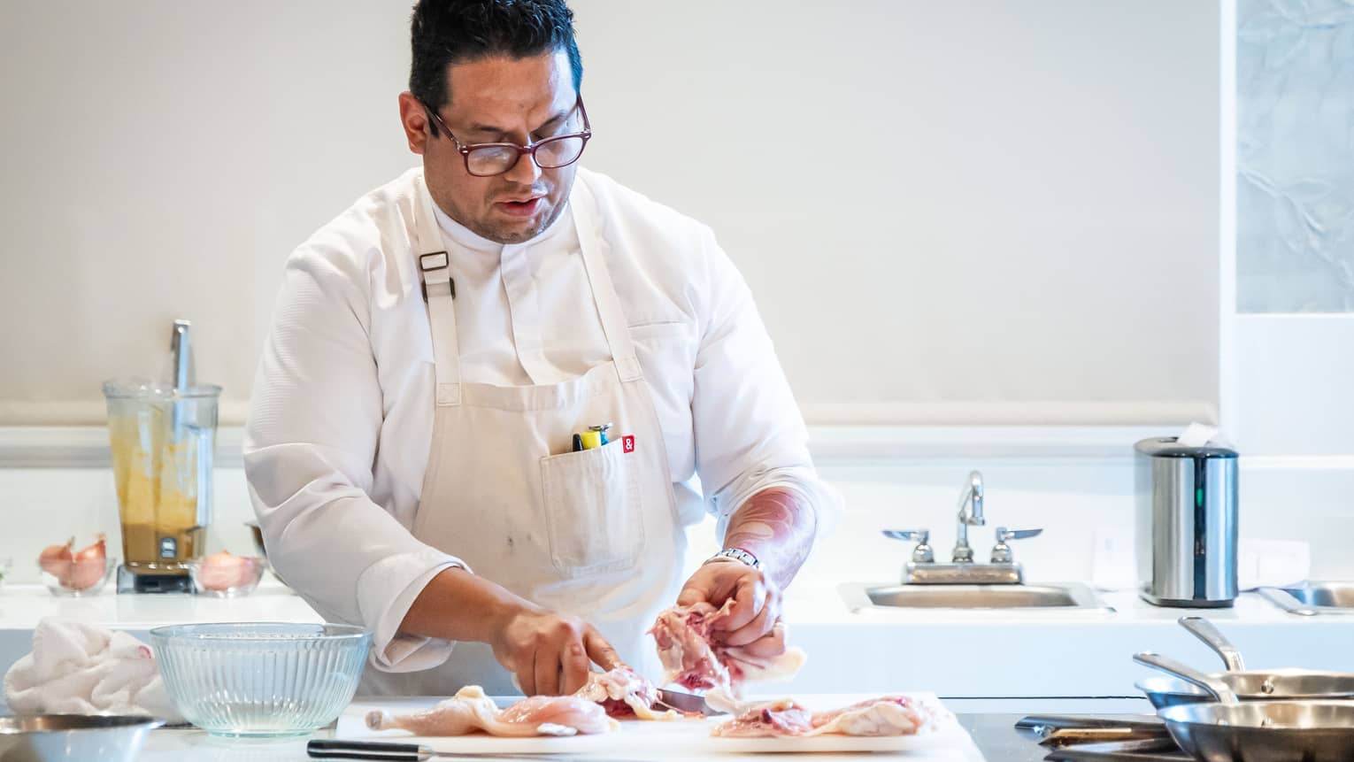 Chef wearing white long-sleeve shirt and a white apron prepares a chicken in a bright kitchen