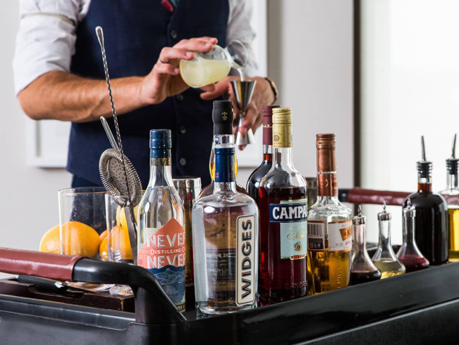 Bartender pours a cocktail at in room cocktail cart with assortment of liquor bottles