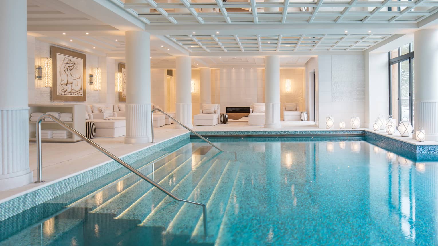 Indoor swimming pool under white beams, pillars, lounge chairs on deck
