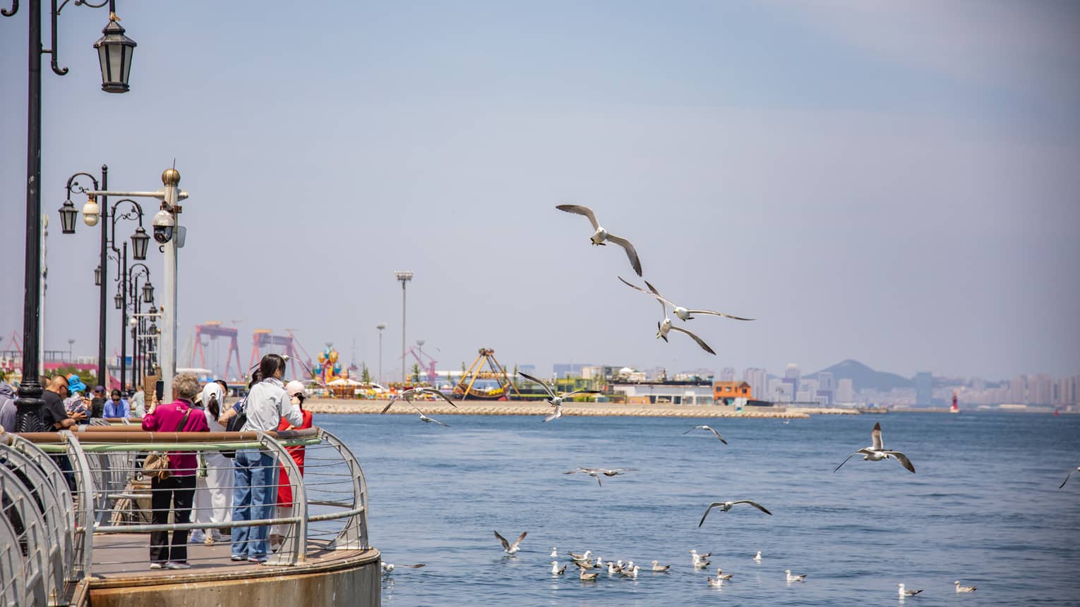 Tourists gather at a sunny pier lined with old-style lamp-posts as seagulls fly nearby, an amusement park in the distance.