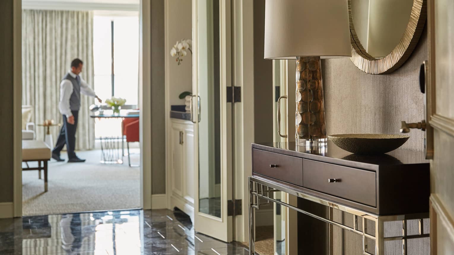 View from hotel suite hall with console table, lamp past doors to staff setting in-room dining table