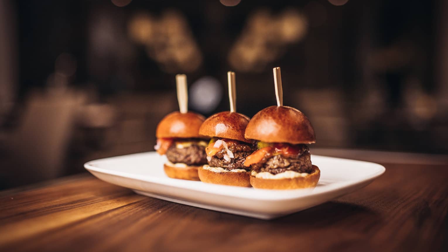 Trio of thick burgers on artisanal buns with wood sticks on white platter
