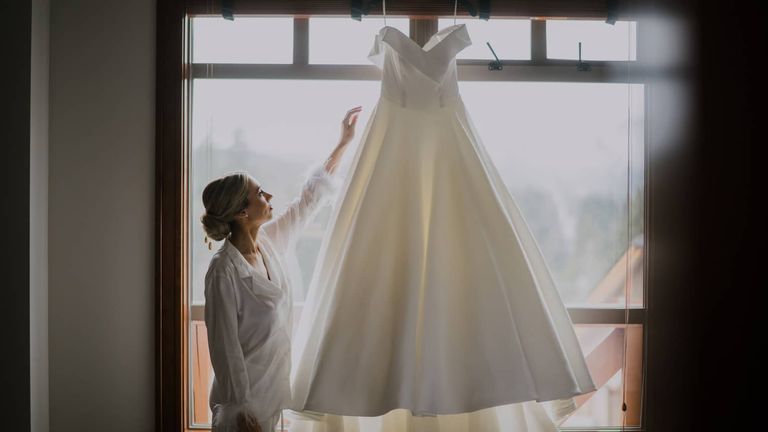 Person reaches hand up to touch wedding dress hanging in front of window