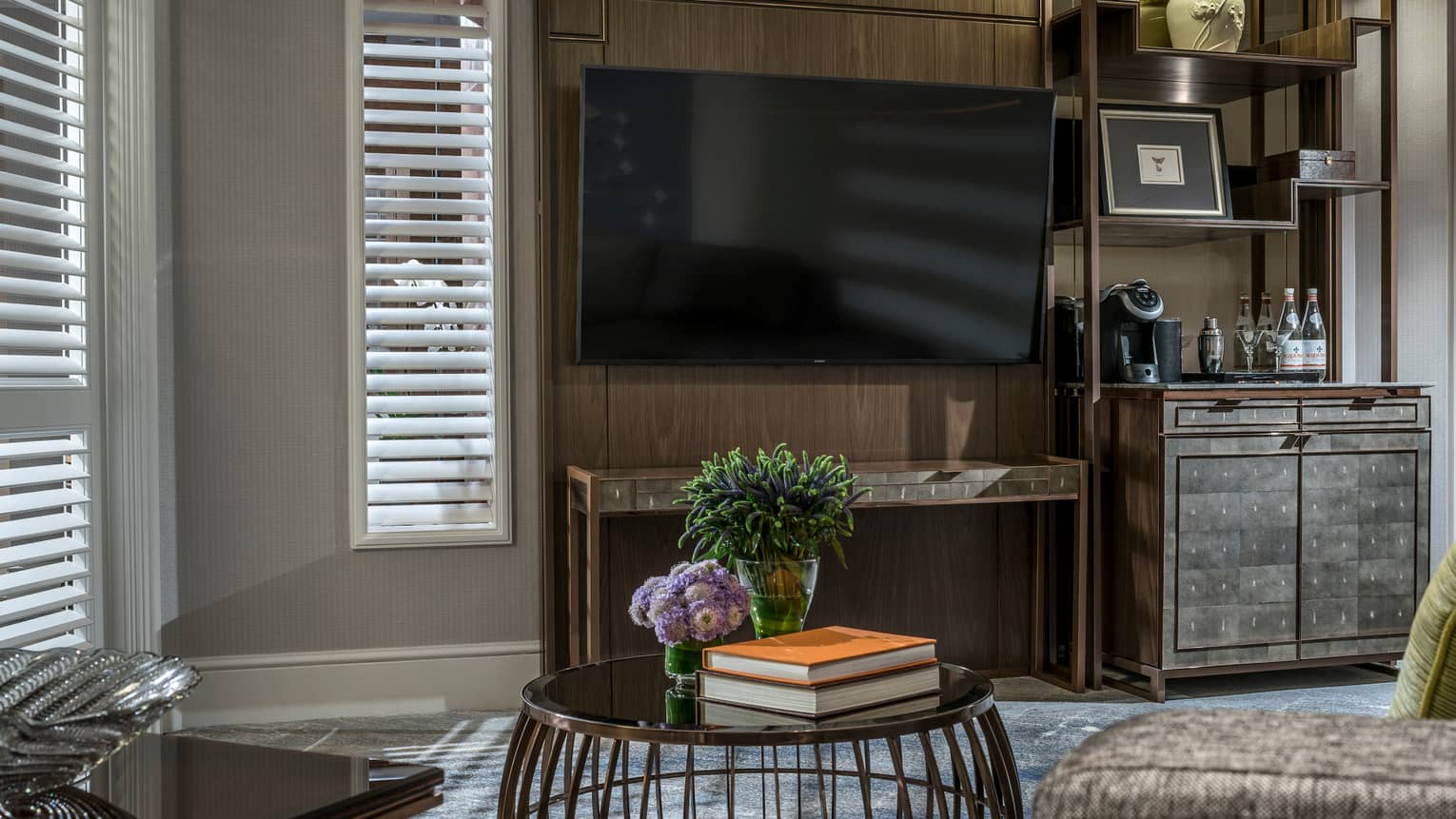A living area featuring a large wall-mounted TV,  sleek wooden cabinet with a coffee machine, and a round table holding flowers and books. Shuttered windows allow soft light to filter in.
