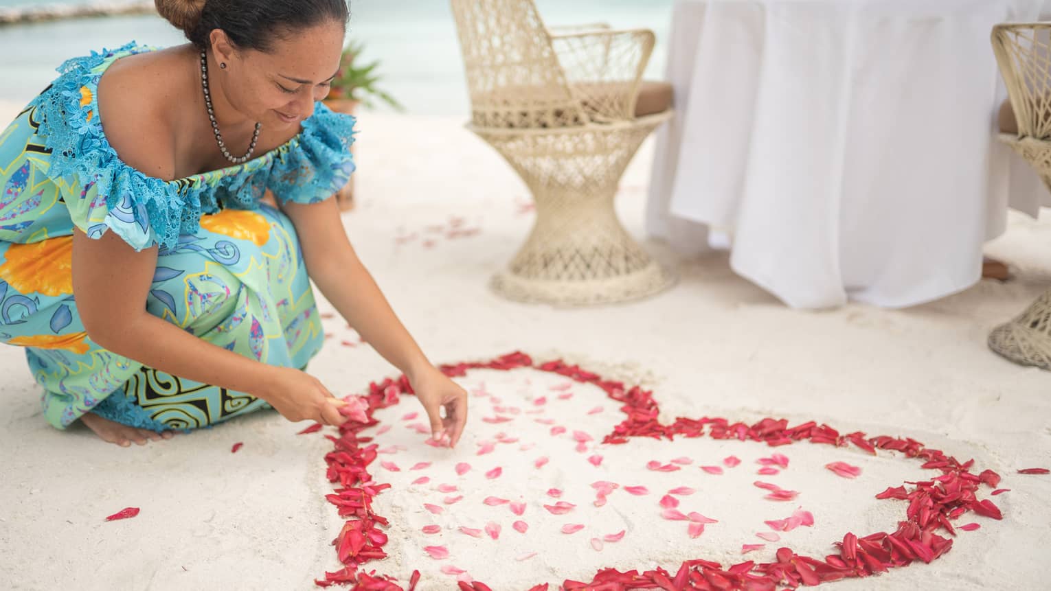 Woman arranges red rose petals into heart shape on sand beach