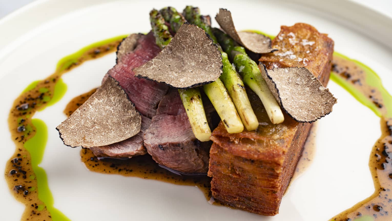Wagyu beef tenderloin and potato Anna topped with four stalks of grilled asparagus with a drizzle of beef jus and basil oil surrounding it all on a white plate